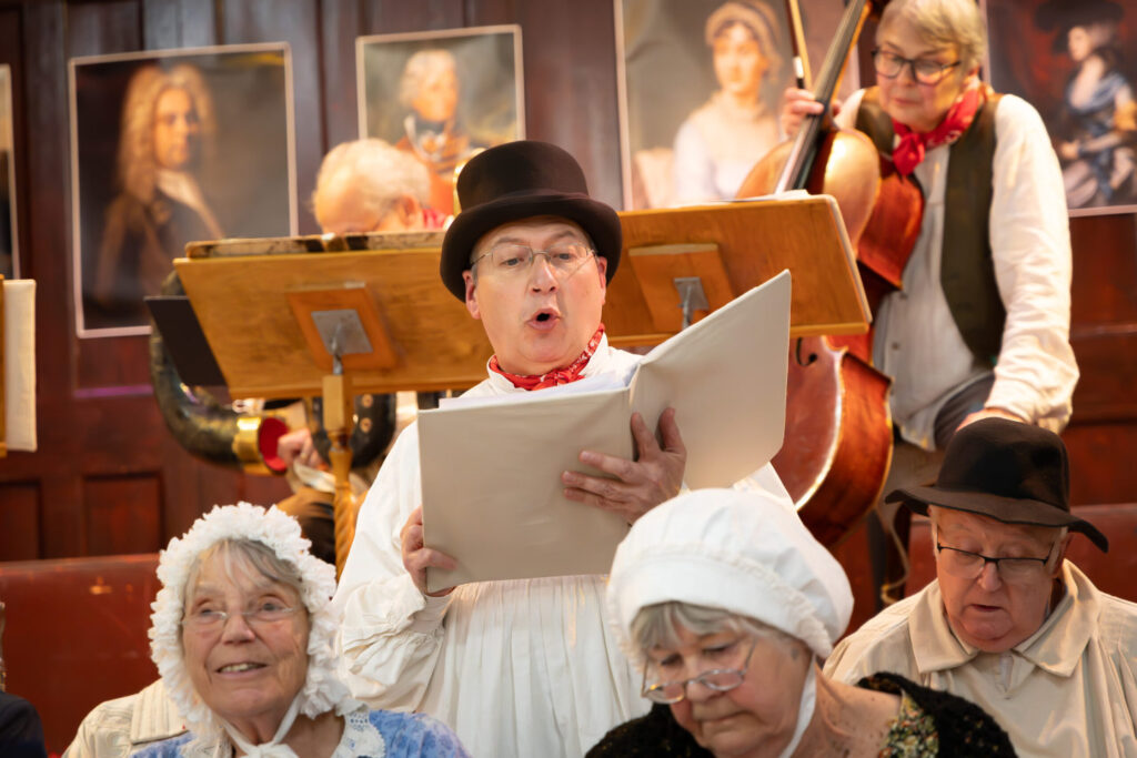 Musical performers on stage, including a male singer in a top hat and a cellist with waistcoat and red, knotted scarf.