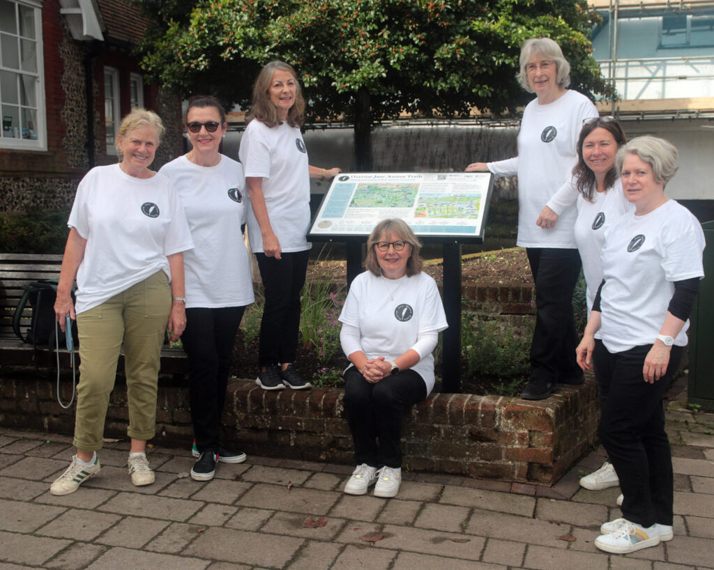 A group of seven ladies wearing white t-shirts with the Jane Austen Trails logo stand at the Trails information board.
