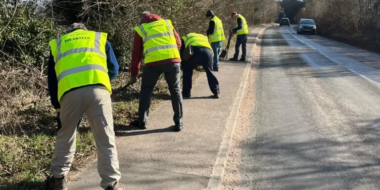 volunteers in hi-vis vests use tools to clear overgrowth from pavement